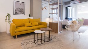 Un salon moderne avec un canapé jaune, une table basse ronde et un fauteuil blanc. On y trouve également une bibliothèque et une plante.