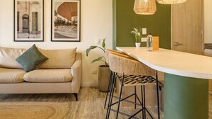 A modern living room with a sofa, a coffee table, and a dining area with a bar stool. There are two framed pictures on the wall.