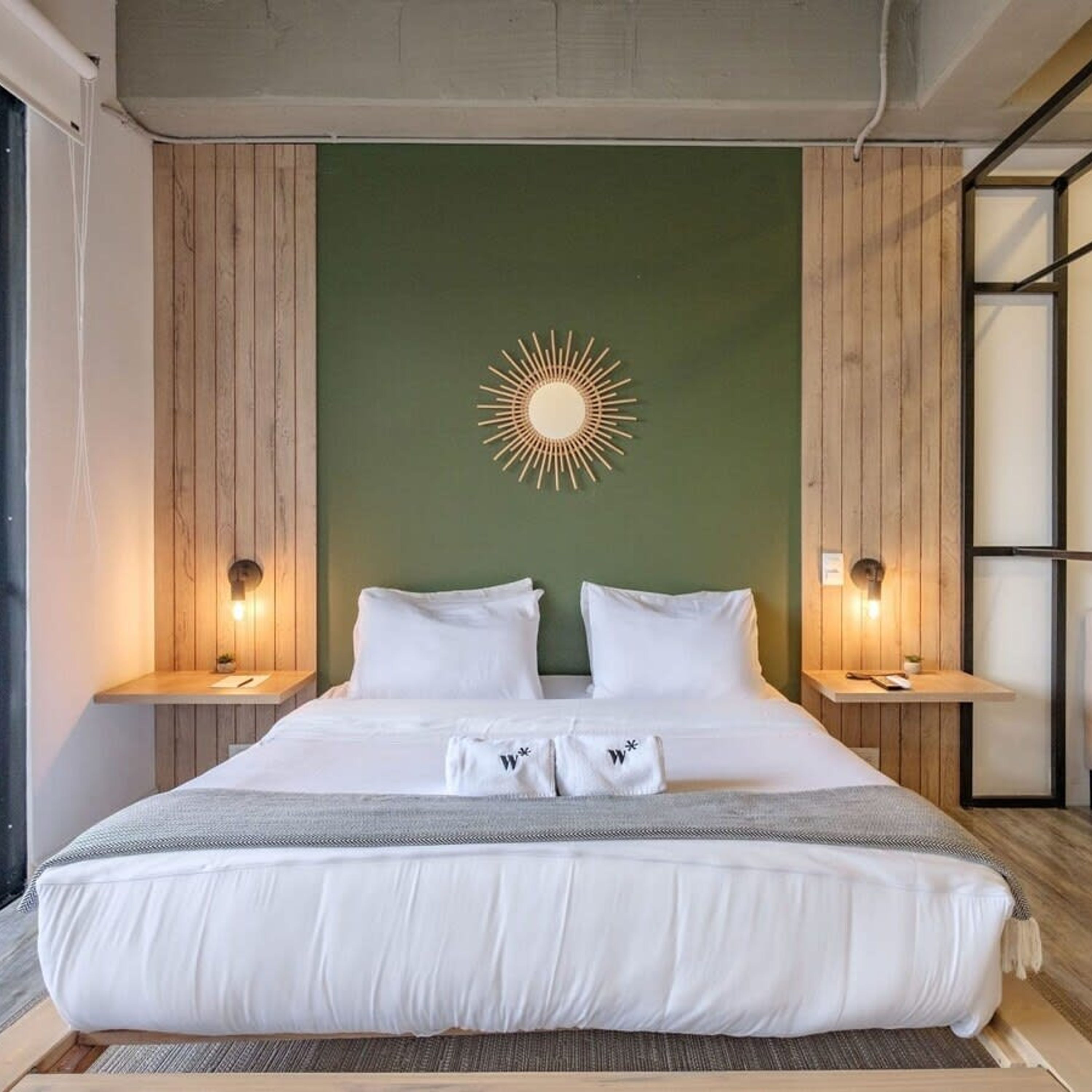 Una habitación de hotel moderna con una cama grande, dos mesitas de noche, una pared de acento verde y una lámpara con forma de sol.