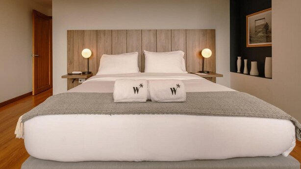 Een moderne hotelkamer met een houten hoofdeinde, twee nachtkastjes met lampen, een groot bed met witte lakens en een ingelijst schilderij aan de muur.