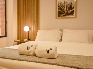 A neatly made bed with two towels featuring a logo, a bedside lamp, and a wall-mounted picture. - w Stylish 2BR in Cabrera (Bogotá)