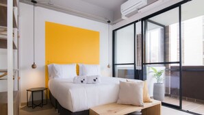 A modern bedroom with a large bed, a bright yellow accent wall, a wooden bench, and a view of the outside through a large window. - w Lovely Loft in Laureles (Medellín)