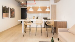 A modern kitchen with white cabinets, a central island with bar stools, pendant lighting, and a visible trash bin. - w Lovely Loft in Laureles (Medellín)