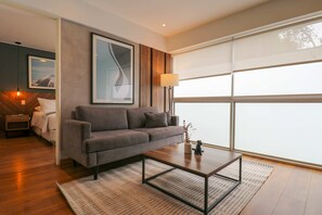A modern living room with a grey sofa, a wooden floor, a coffee table, and a large window with blinds. - w Dreamy 1BR in Barranco (Lima)