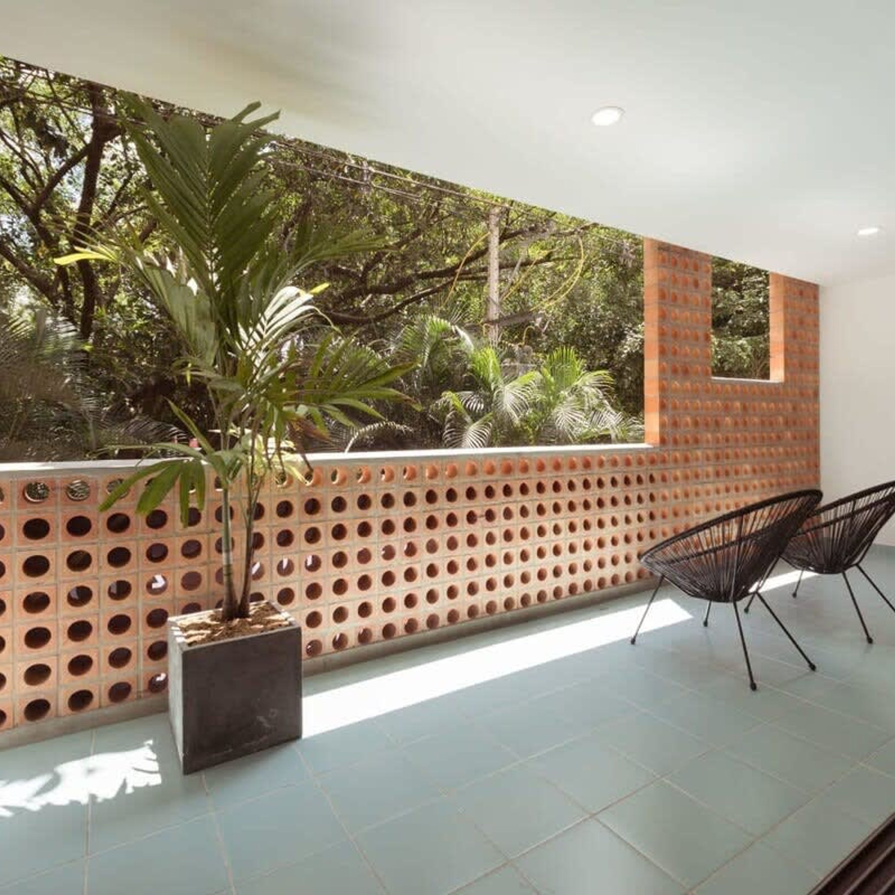 Un patio extérieur moderne avec un mur en briques perforées, un pot de fleurs et deux chaises noires en fil de fer.