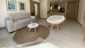 Un salon avec un canapé, une table basse ronde et un fauteuil en osier. On aperçoit une cuisine au fond de la pièce.