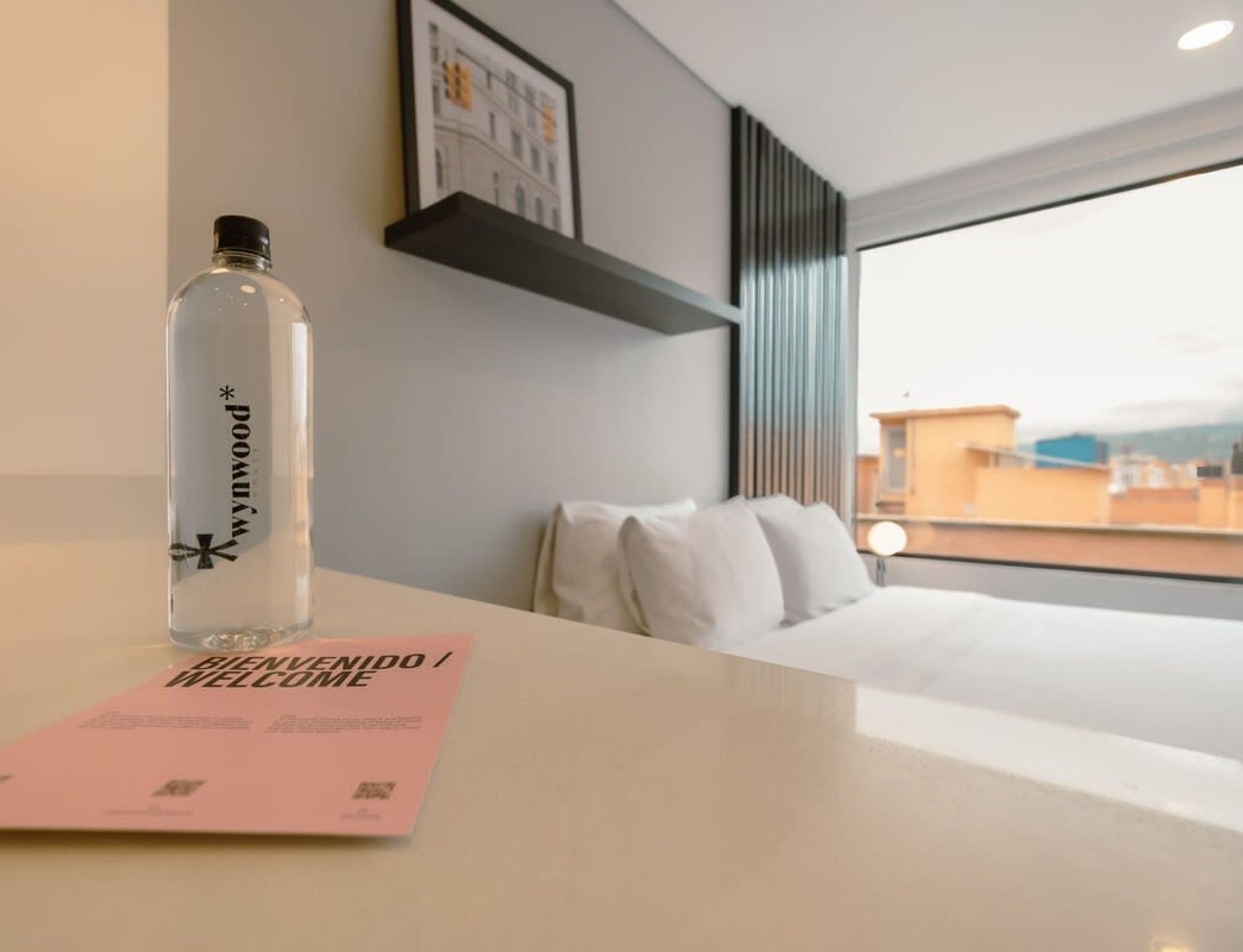 A hotel room with a bed, a bedside table with a water bottle and a welcome card, a window with a view of buildings, and a framed picture on the wall.