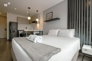 A modern hotel room with a large bed, a small kitchenette, and a view of the city through the window.