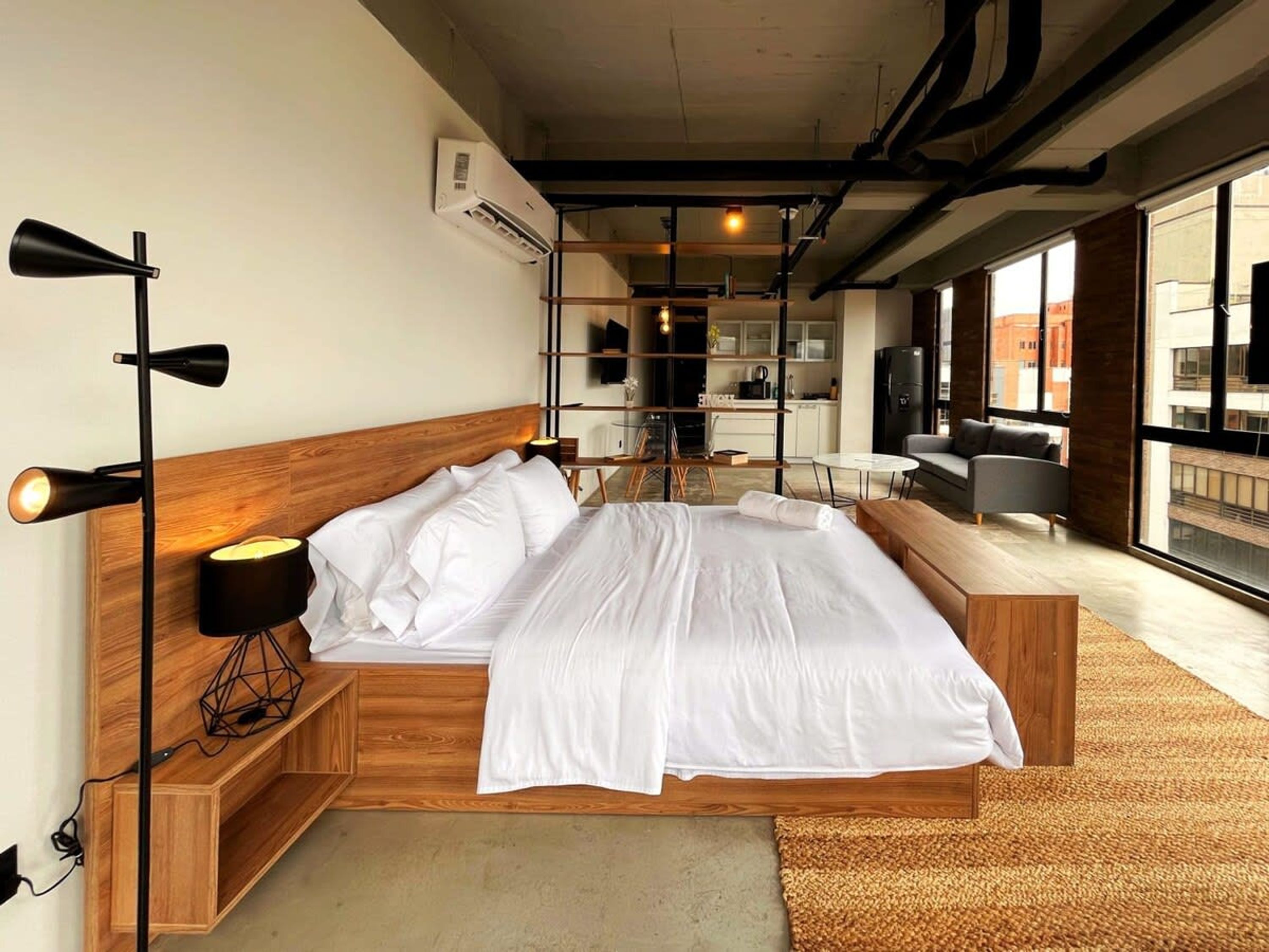 A modern hotel room with a large bed, wooden headboard, bedside lamps, a small wooden side table, a large window with a city view, and a kitchenette in the background.