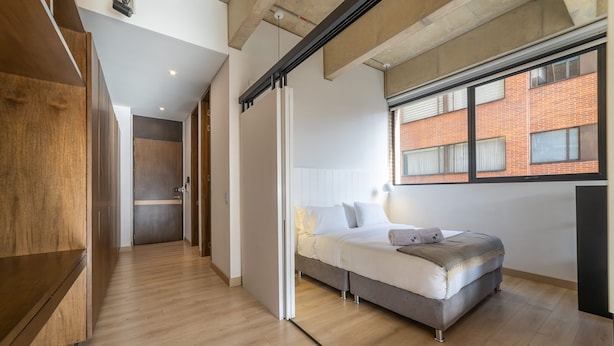 A modern bedroom with a bed, wooden flooring, a window with a view of a brick building, and a sliding door. - Splendid Loft in Parque 93 (Bogotá)