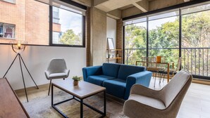 A modern living room with a blue sofa, a wooden coffee table, and a grey armchair. There is a large window providing a view of trees and a brick building. - Splendid Loft in Parque 93 (Bogotá)