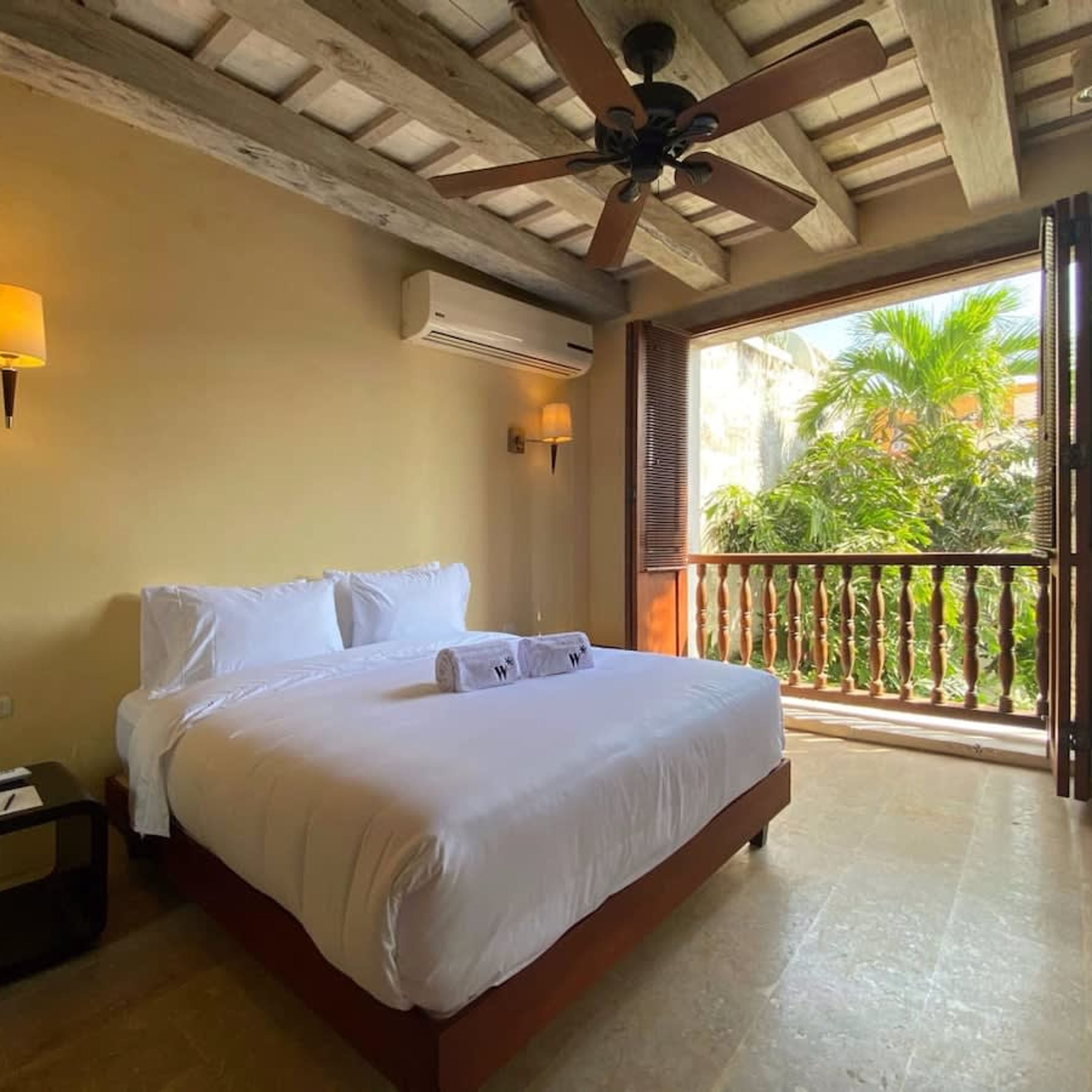 A hotel room with a large bed, an air conditioning unit, a ceiling fan, a nightstand, and a view of palm trees through a balcony door.