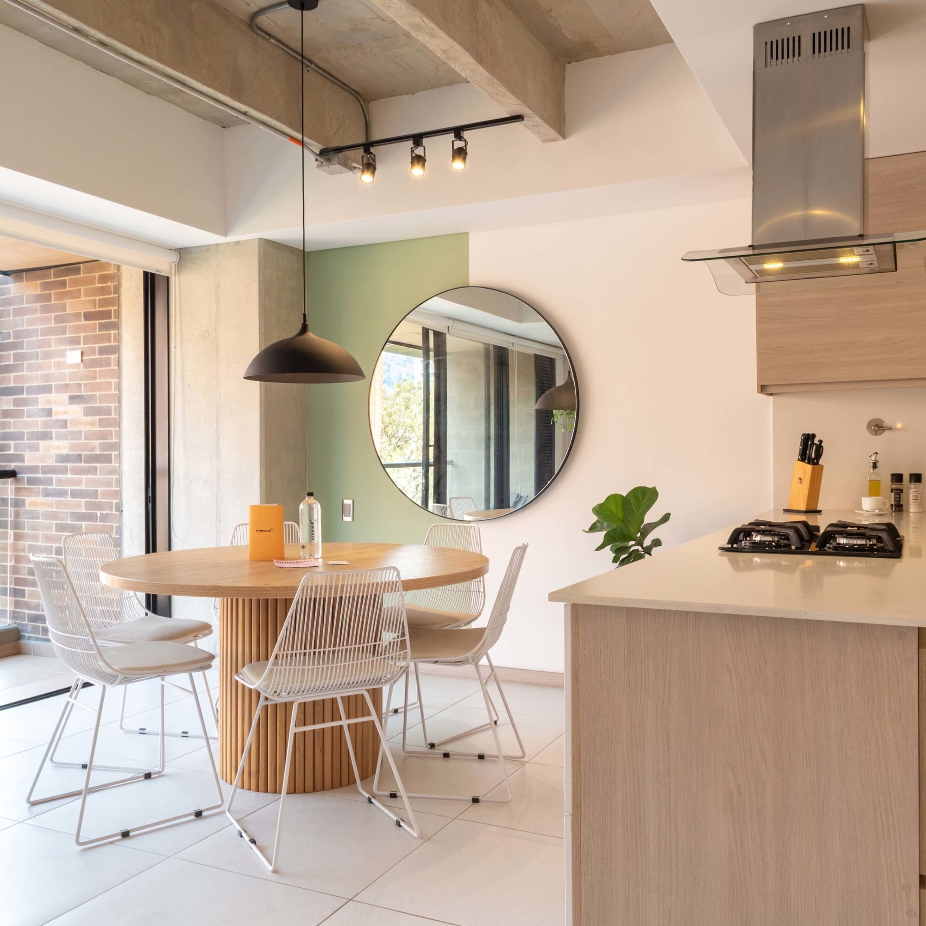 A modern kitchen with a round dining table, white chairs, a hanging pendant light, and a large mirror on the wall.