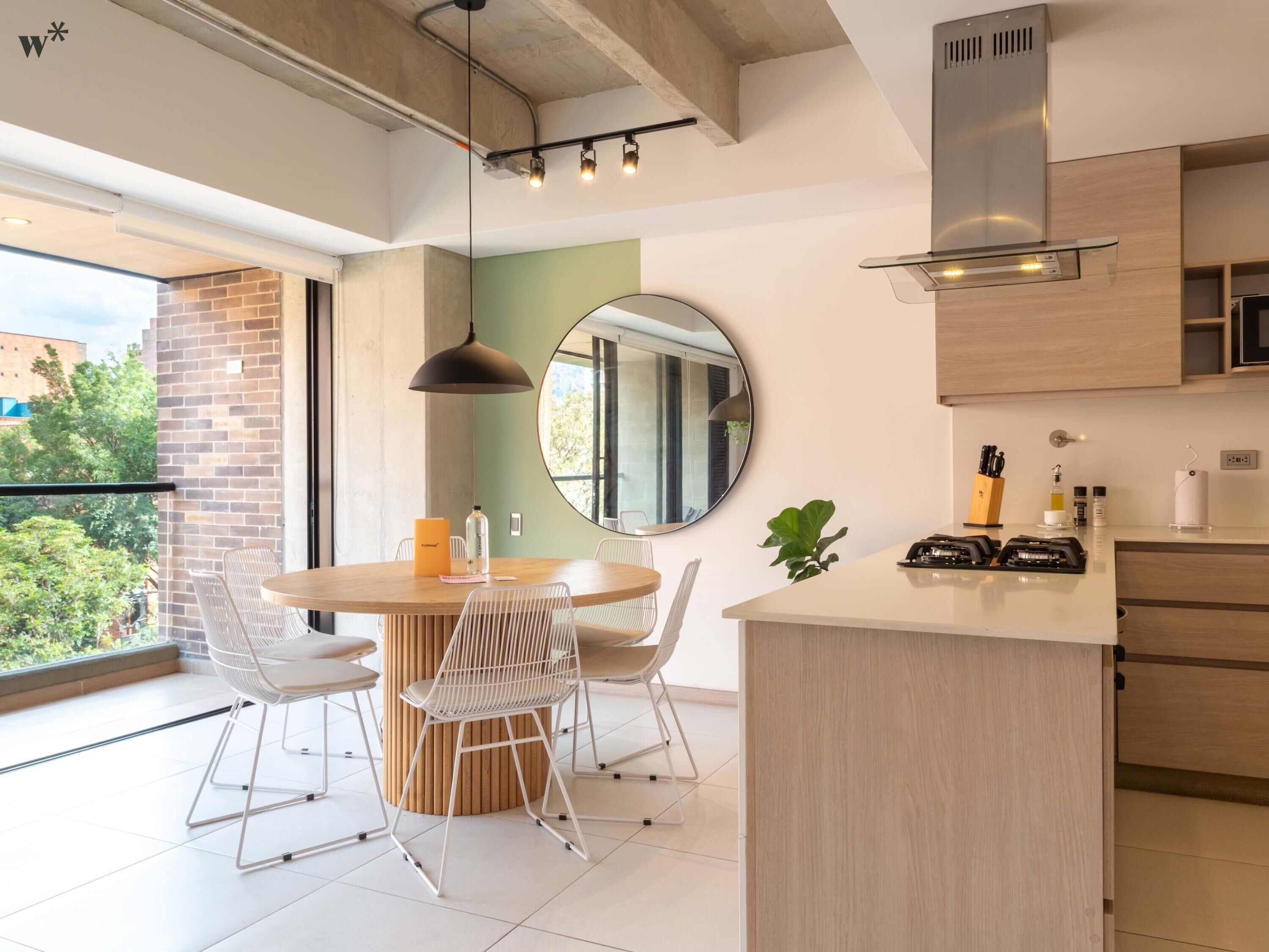 A modern kitchen with a round dining table, white chairs, a hanging pendant light, and a large window overlooking greenery.