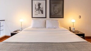Una cama bien hecha con sábanas blancas, dos mesitas de noche con lámparas y dos cuadros en la pared.