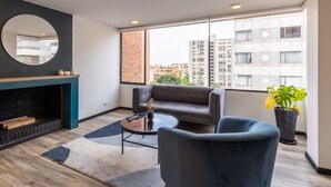A modern living room with a fireplace, a round mirror, a grey sofa, a coffee table, and a large window with a city view.