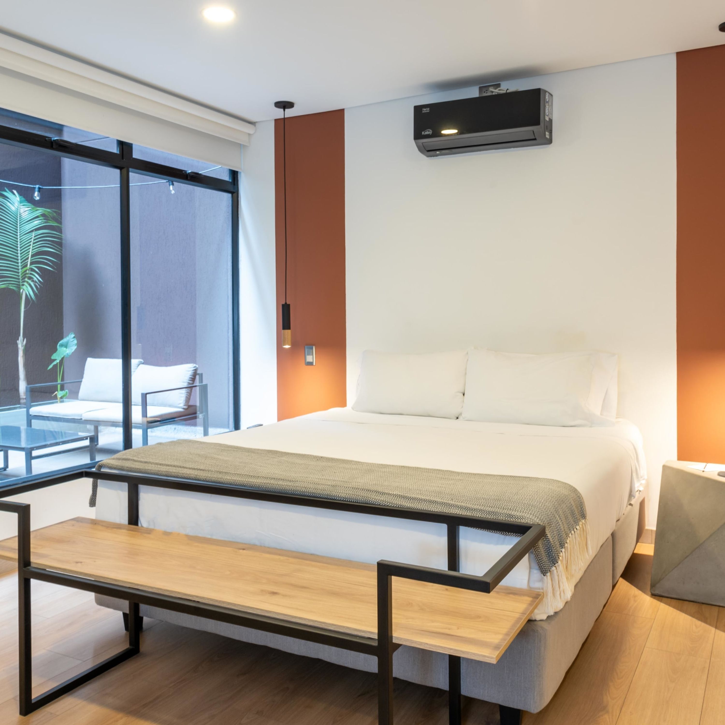 A modern bedroom with a large bed, a bench at the foot of the bed, a window with a view of a balcony, and an air conditioning unit on the wall.