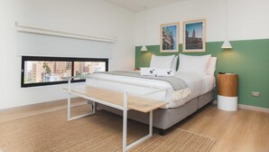 A modern bedroom with a bed, a bench, a window with a view, and two framed pictures on the wall. - Jungle Style 1BR in Laureles (Medellín)