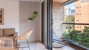 Un salon moderne avec un canapé, deux fauteuils, une plante suspendue et une grande fenêtre donnant sur un paysage urbain.