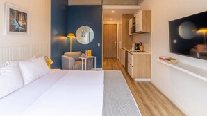 Une chambre d’hôtel moderne équipée d’un grand lit, d’un bureau, d’une chaise, d’une petite table, d’un miroir, d’une télévision et d’une kitchenette.