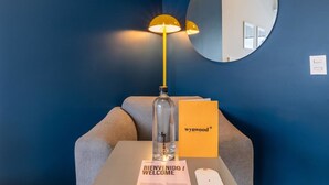 Un espace d’accueil d’hôtel avec un mur bleu, un miroir rond, une lampe, une bouteille, une carte de bienvenue et une petite table.