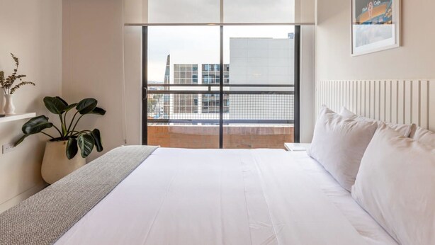 Une chambre avec un grand lit, une vue sur les immeubles à travers une porte vitrée, une plante en pot et un tableau encadré au mur.