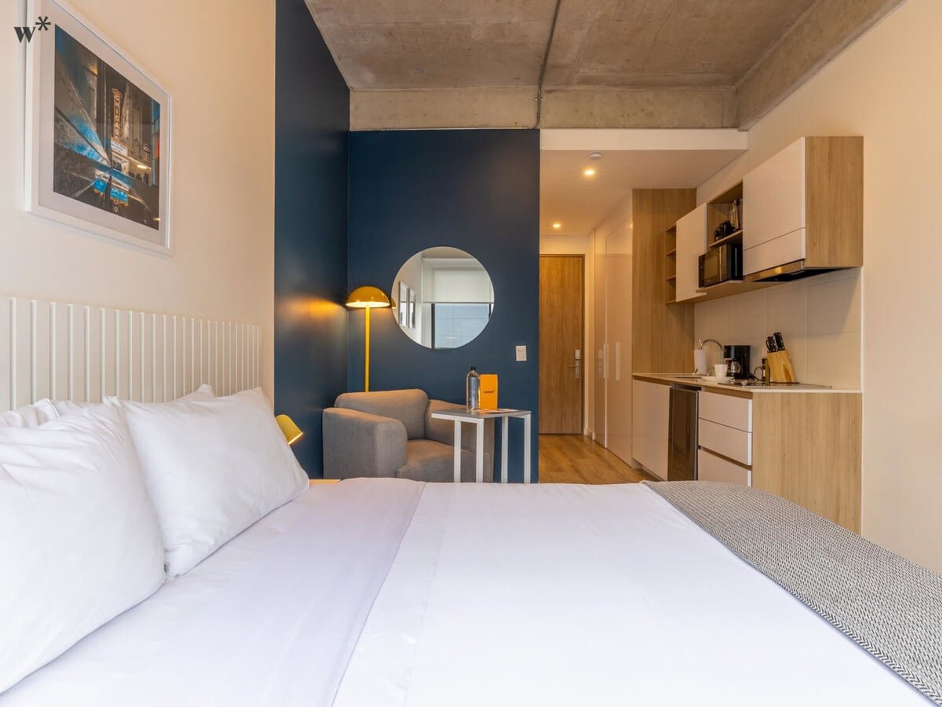 A modern hotel room with a bed, a desk, a chair, a round mirror, and a kitchenette.