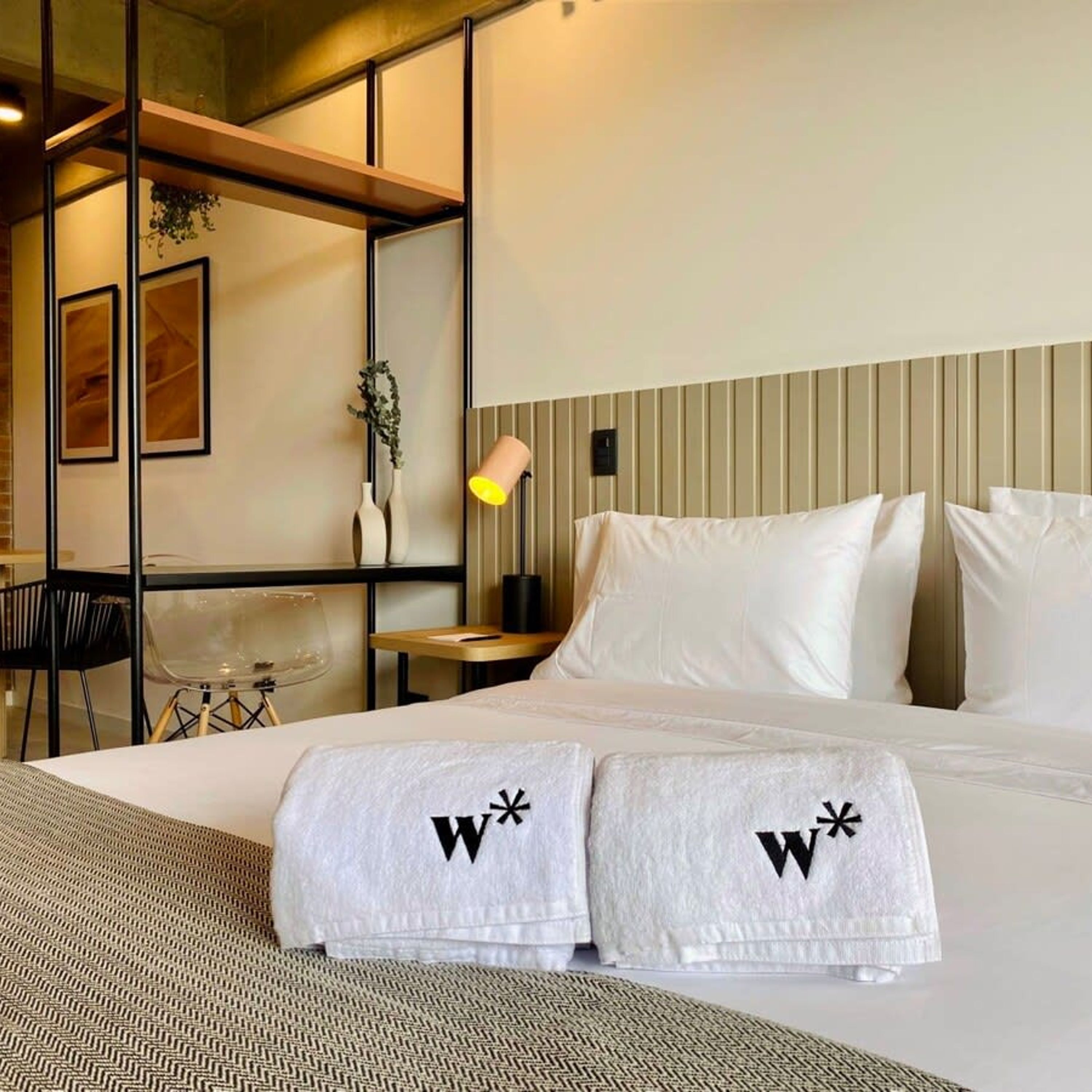 Habitación de hotel con cama, dos toallas blancas con un logotipo, escritorio, silla, lámpara y planta.