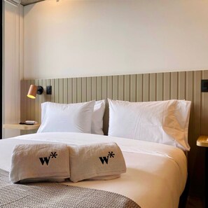 A hotel room with a bed, pillows, a bedside table, a lamp, and a wall-mounted light.