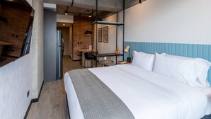 A modern hotel room with a large bed, a desk with a chair, a small table, and a kitchenette in the background.