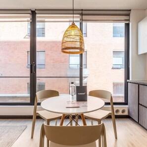A modern dining area with a round table, four chairs, a wicker pendant light, and a view of a brick building through large windows.