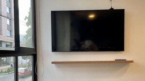 A modern room with a large flat-screen TV mounted on the wall, a small wooden shelf below it, a round black table, and a large window providing a view of the street outside. - w Extraordinary 2BR in Laureles (Medellín)