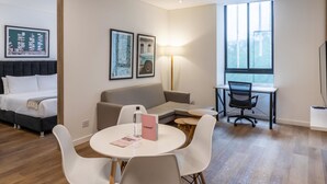 Ein modernes Hotelzimmer mit Bett, Sofa, Esstisch mit Stühlen und Schreibtisch mit Stuhl. An der Wand befinden sich gerahmte Bilder, eine Lampe und ein Fenster mit Blick auf Gebäude.
