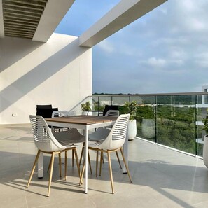 Un espace de restauration extérieur moderne, avec une table en bois et des chaises blanches, entouré d’une rambarde en verre donnant sur un panorama urbain.