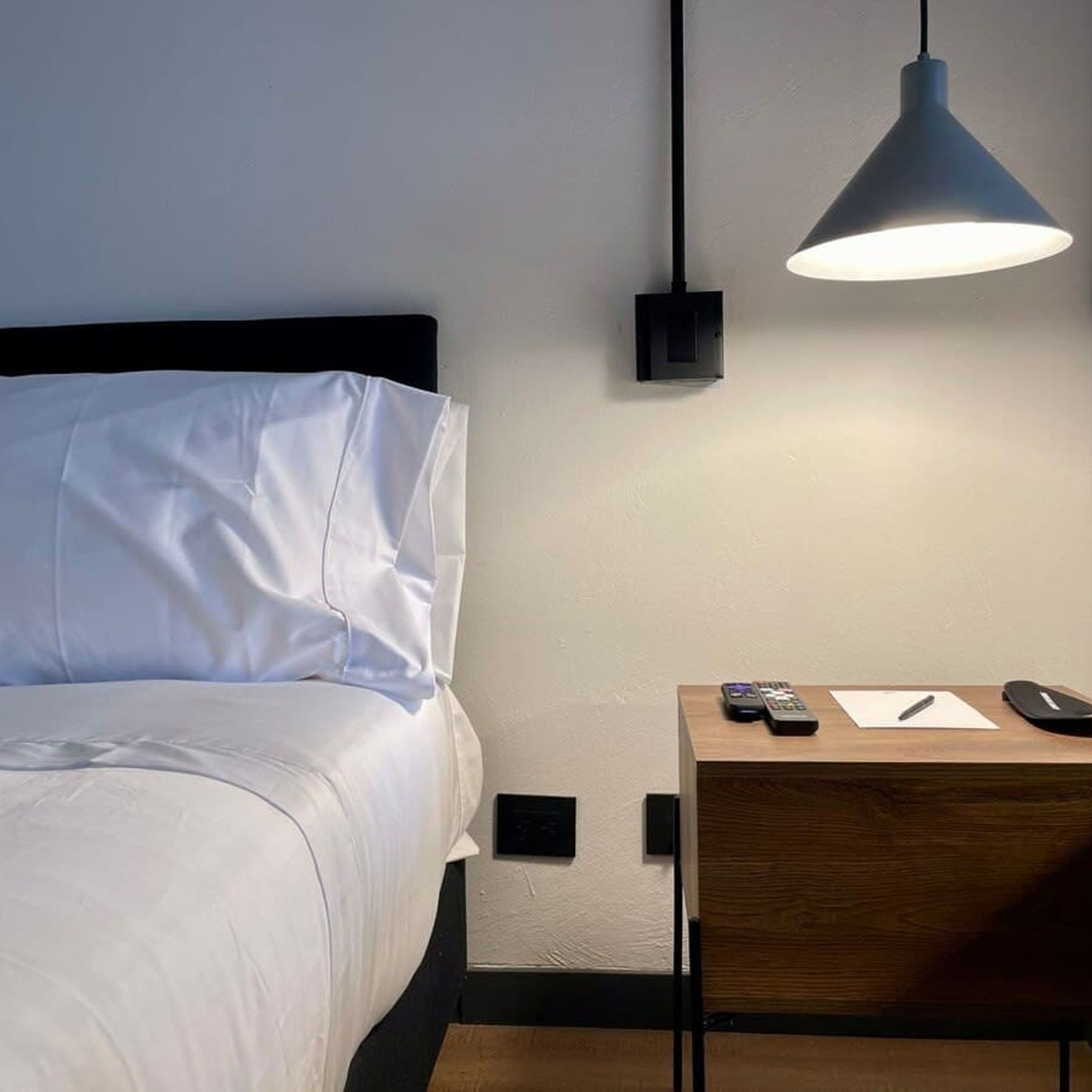 Un lit bien fait, avec du linge de lit blanc, une table de chevet avec une lampe et une télécommande posée sur la table.