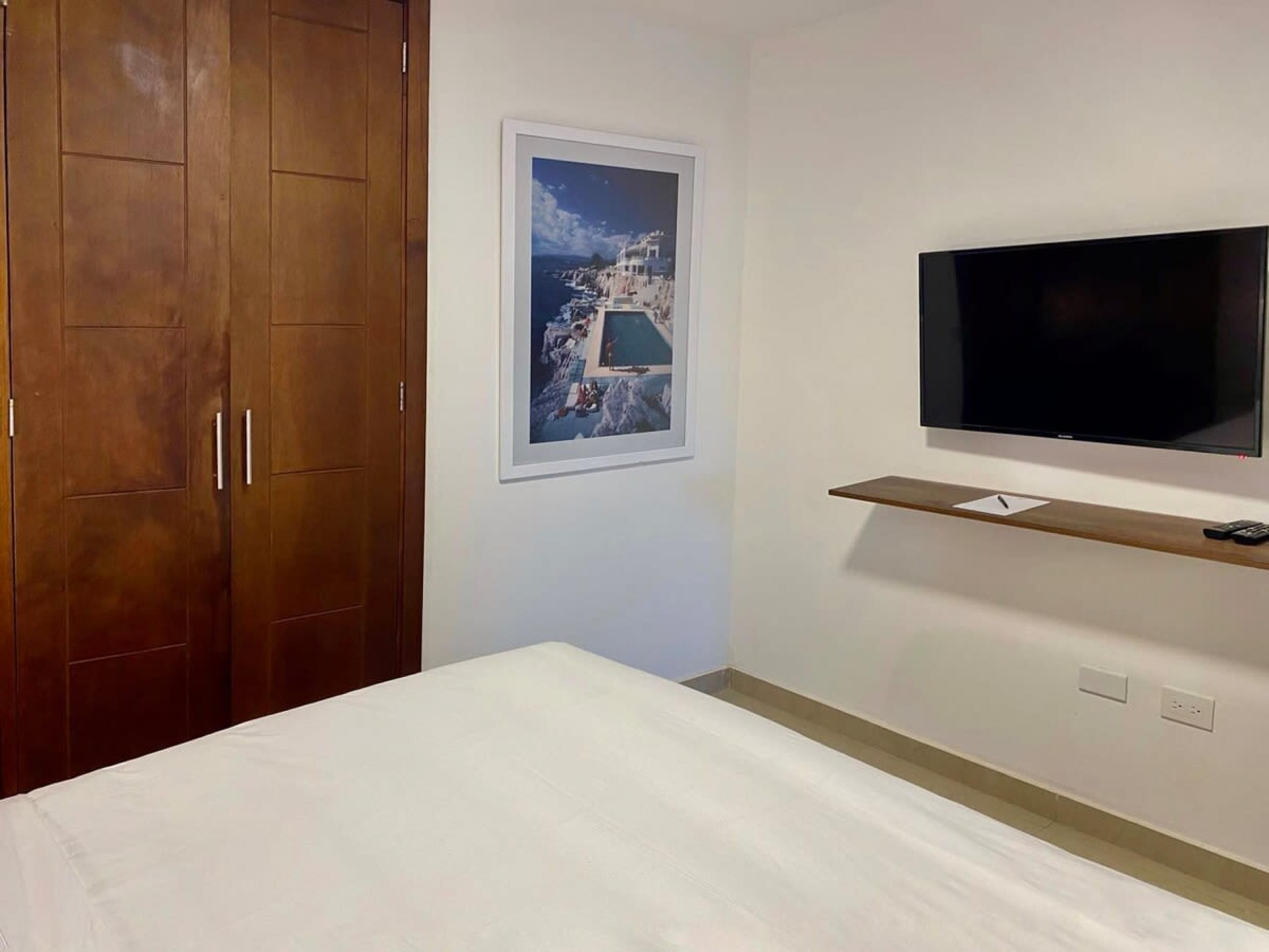 Un dormitorio con una cama, un armario de madera, un cuadro enmarcado en la pared y un televisor montado.