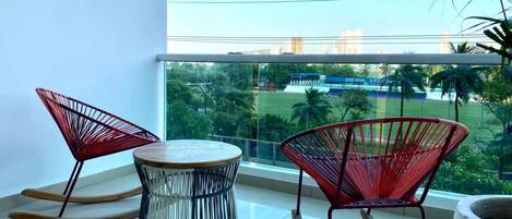 A balcony with two rocking chairs and a round table, overlooking a sports field and buildings.