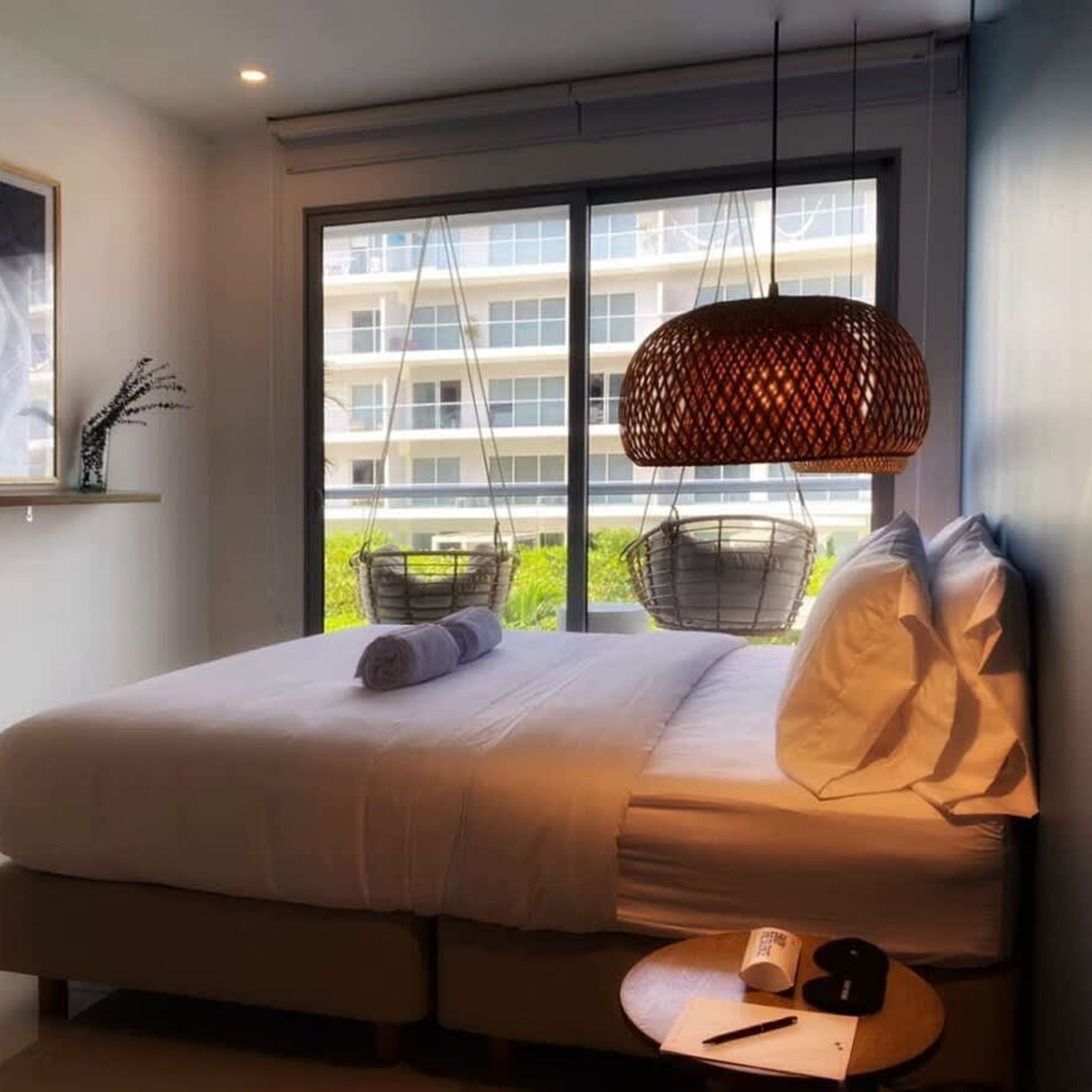 A modern bedroom with a large bed, a hanging light fixture, and a view of a building through the window.