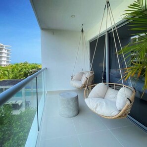 A balcony with two wicker swing chairs, a round marble stool, and a glass railing overlooking a building and greenery. - w Fantastic 2BR w AC in Sunny Cartagena (Cartagena)