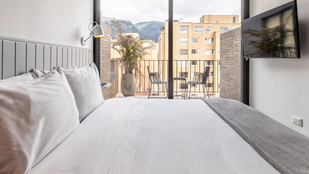 A modern hotel room with a large bed, a balcony with a view of buildings and mountains, a mounted TV, and a potted plant. - Amazing Loft With Balcony in El Nogal (Bogotá)