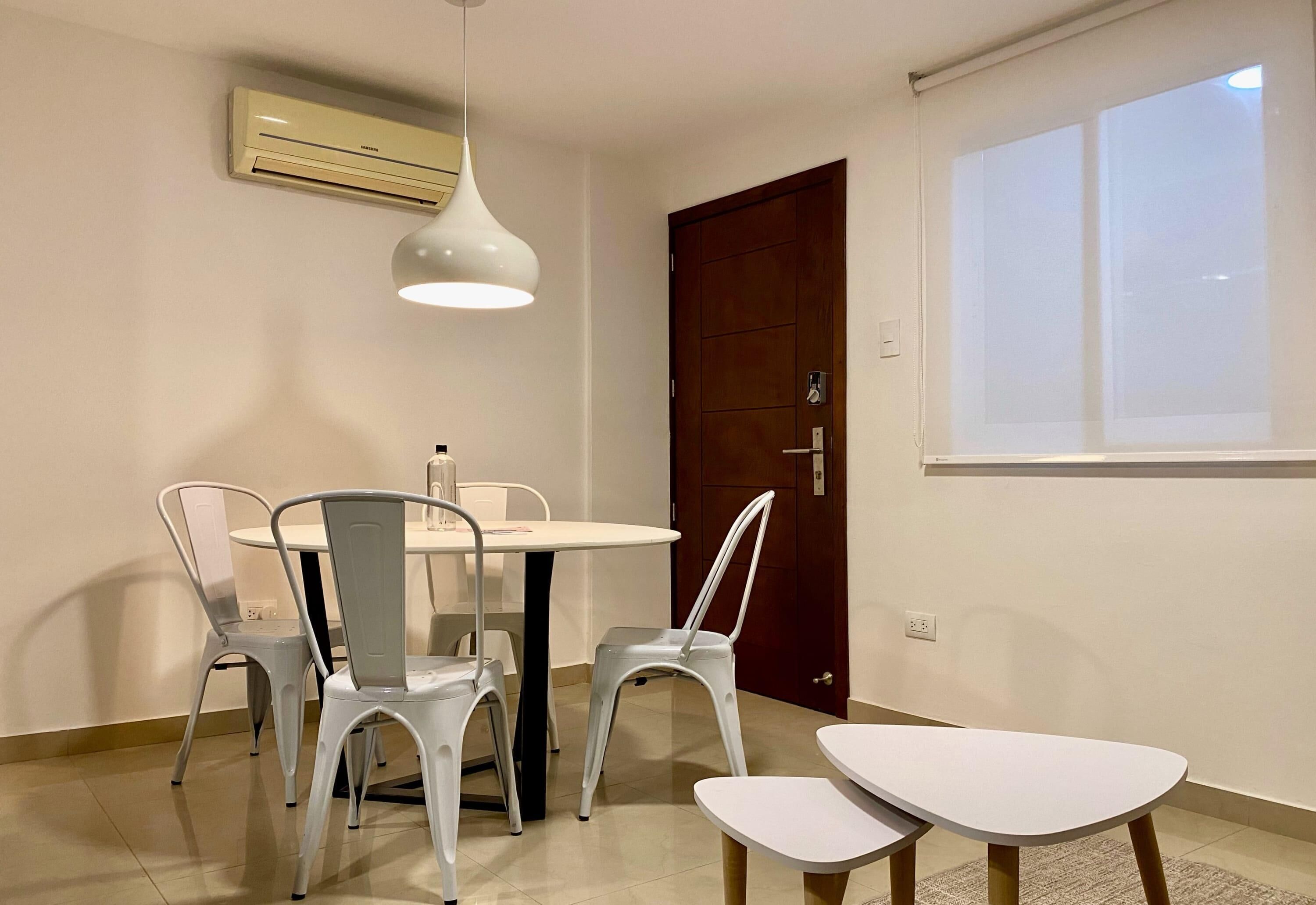 Un comedor con una mesa redonda y cuatro sillas, una lámpara colgante blanca, un equipo de aire acondicionado, una puerta de madera y una ventana con persianas.