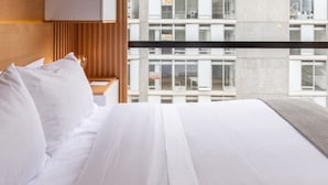 Una habitación de hotel moderna con una cama grande, cabecera de madera y vistas a un rascacielos a través de ventanas del piso al techo.
