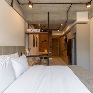 Une chambre d’hôtel moderne dotée d’un grand lit, d’une paroi en verre séparant la chambre à coucher de la cuisine, et d’un coin repas avec une table et des chaises.