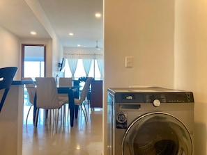 Une buanderie équipée d’un lave-linge et d’un sèche-linge à chargement frontal, un coin repas avec une table et des chaises, et une fenêtre avec des rideaux.