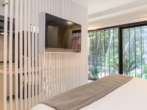 Un dormitorio moderno con un televisor montado en un panel blanco, una cama con una colcha estampada y un ventanal con vistas a la vegetación.