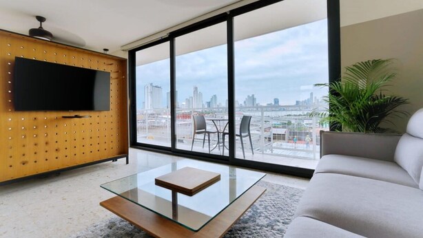 Un salon moderne avec une table basse en verre, un canapé et un téléviseur fixé à un mur en bois. De grandes fenêtres offrent une vue sur la ville.