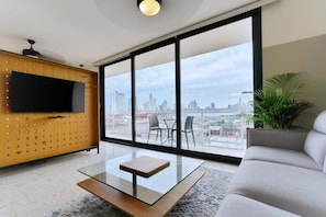 A modern living room with a glass coffee table, a sofa, and a TV mounted on a wooden wall. Large windows offer a city view. - w Refined 1BR in Casco Antiguo (Panama City)
