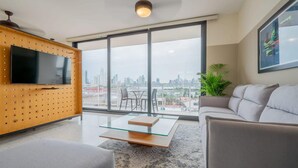 Un salon moderne avec un grand téléviseur, une table basse en verre et un canapé. De grandes fenêtres offrent une vue sur la ville.