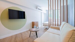A modern living room with a flat-screen TV mounted on a wall, a round table with a bottle and a notepad, a sofa, and a window with vertical blinds. - Amazing Loft in Casco Viejo (Panama City)