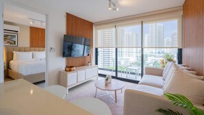 Une chambre d’hôtel moderne équipée d’un lit, d’une télévision, d’un canapé, d’une table basse et d’une grande fenêtre donnant sur la ville.